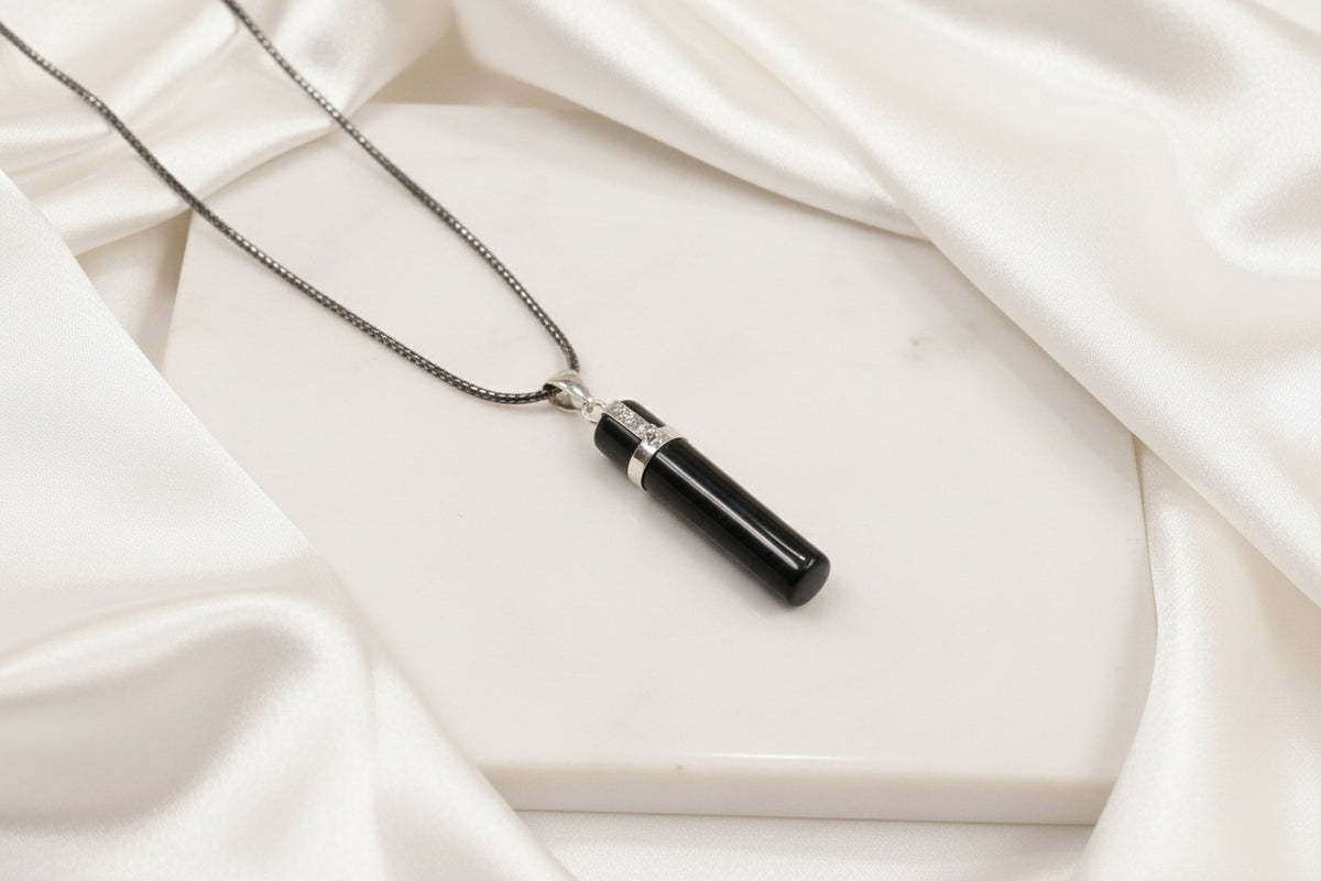 A black cylindrical pendant necklace with polished silver metal accents and a silver-toned chain, displayed on a textured dark gray surface.