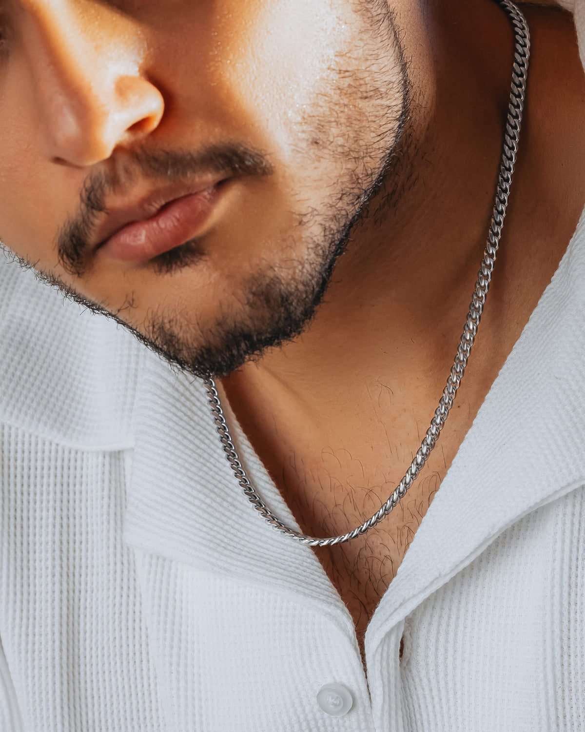 A close-up of a man wearing a silver-toned chain necklace over a white textured collared shirt.