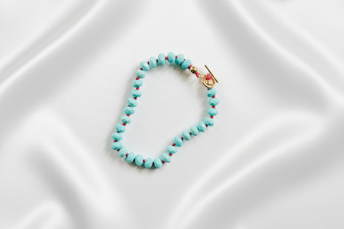 A beaded bracelet made of small turquoise-blue beads with tiny red accents, arranged in a loop with a gold-toned toggle clasp, photographed on a clean light gray background.