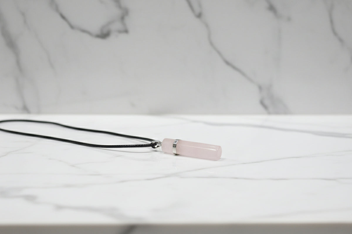 A rose quartz crystal pendant necklace on a black cord, displayed on a textured gray background.