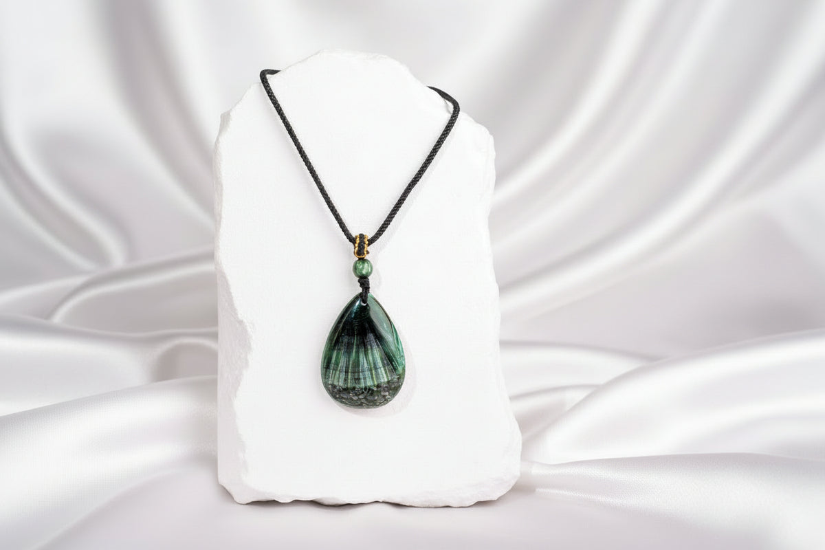 A teardrop-shaped green stone pendant necklace with black and white banded striations, hanging from a black cord with a small green bead and gold spacer, displayed on a white stone block background