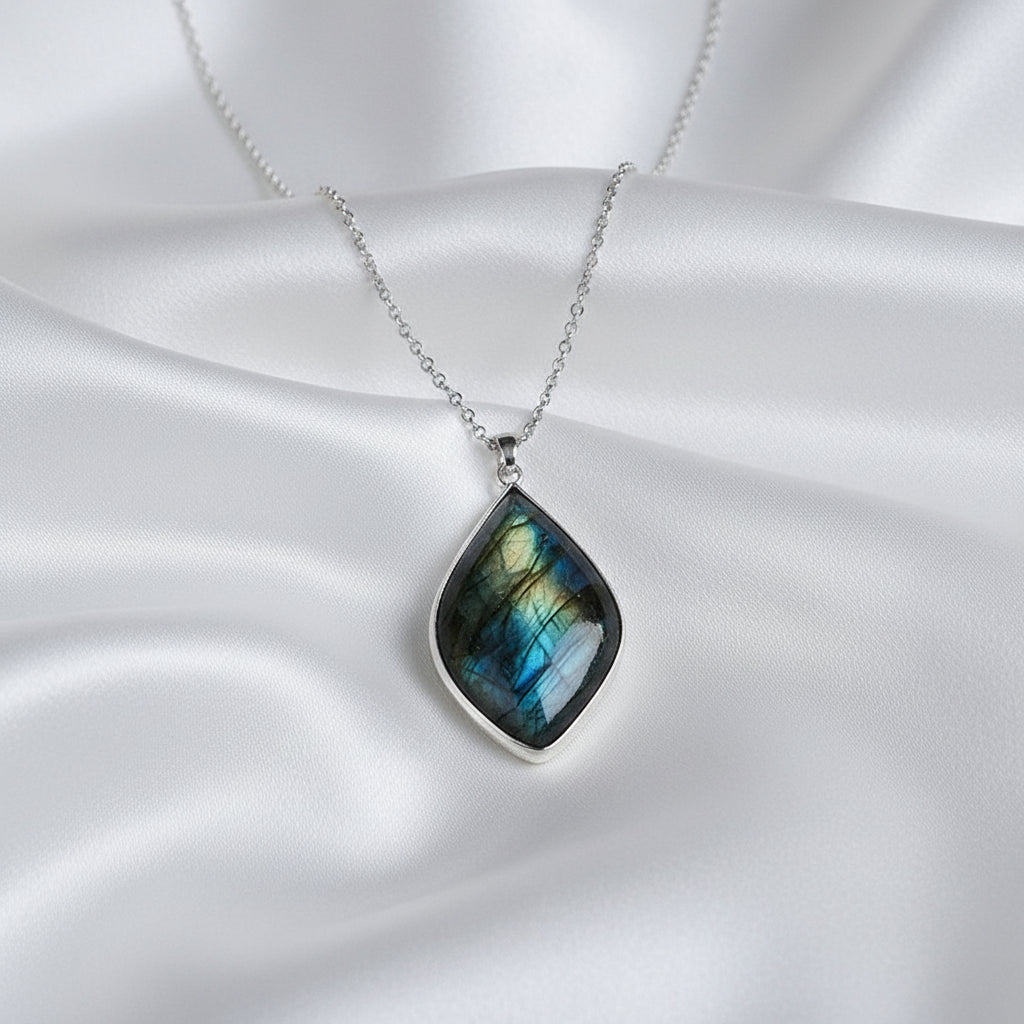 A teardrop-shaped labradorite pendant necklace with a silver-toned chain, displayed on a silky white surface.