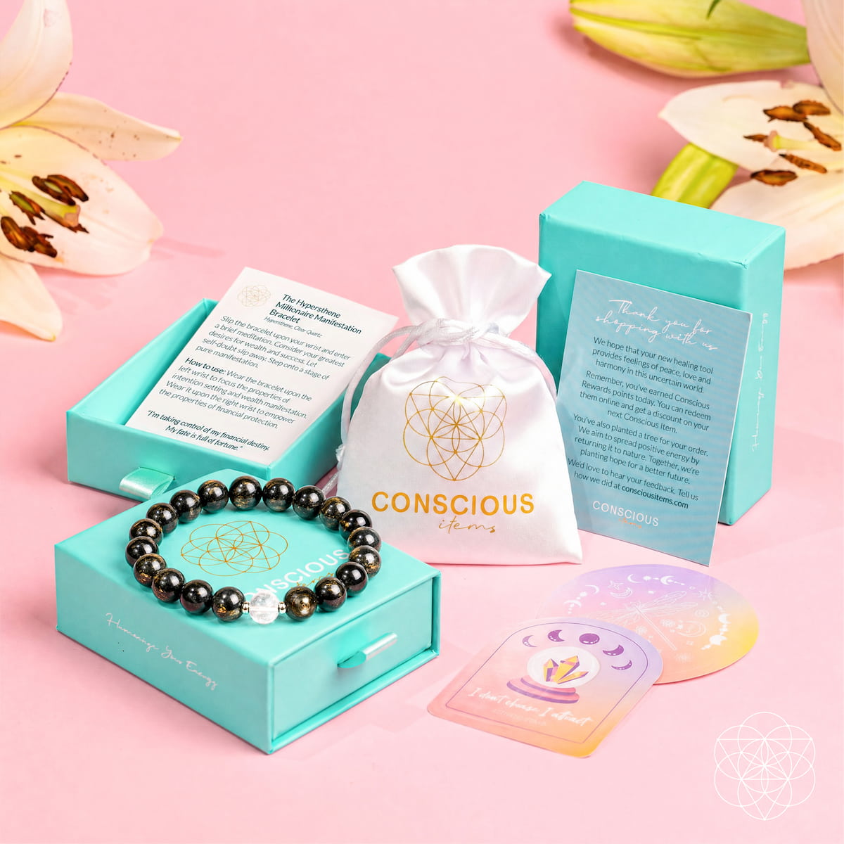 A jewelry gift set on a pink background, featuring a black bead bracelet in a turquoise box, a white drawstring pouch with 'CONSCIOUS lives' printed on it, a turquoise card, and two iridescent rose quartz healing crystals.
