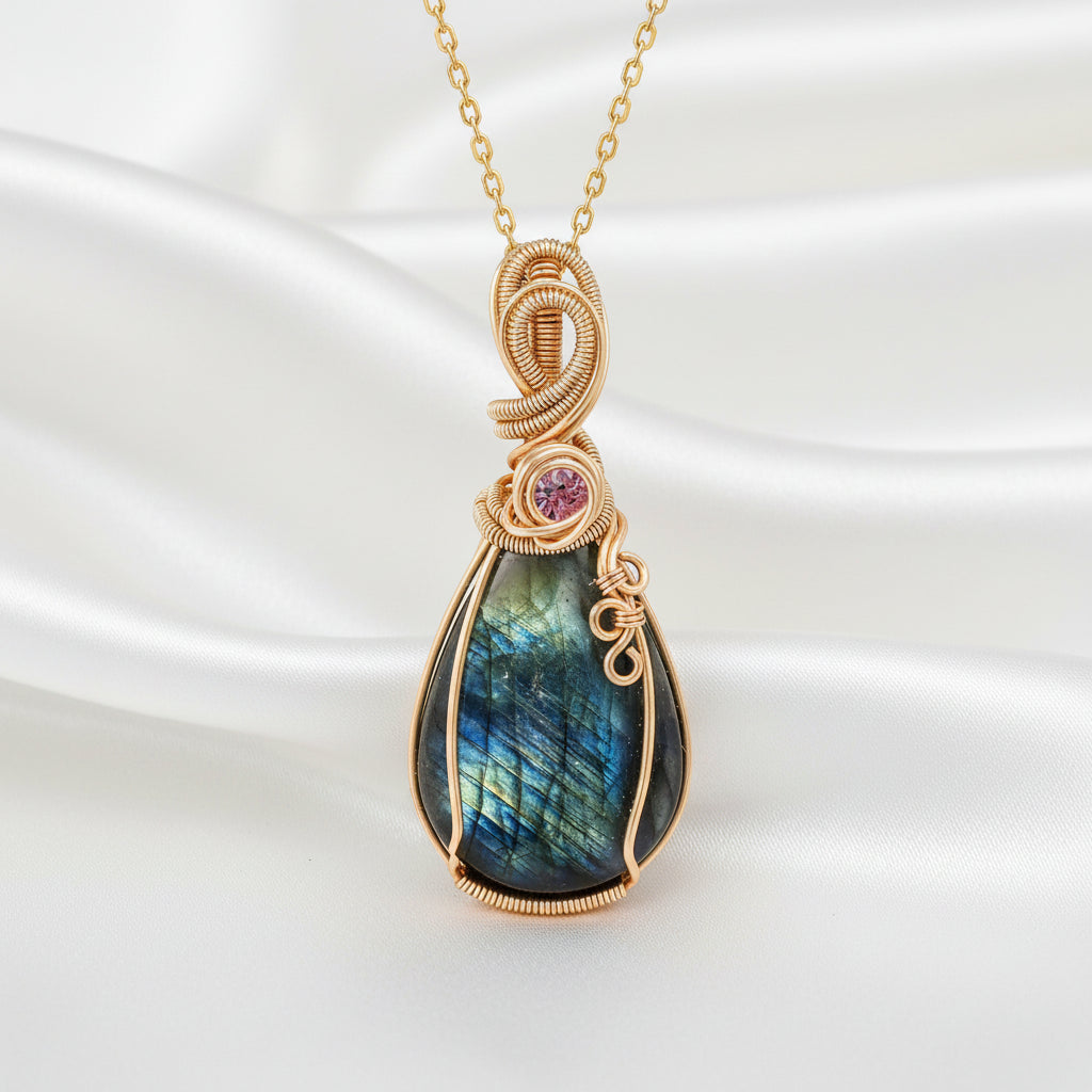 A teardrop-shaped labradorite pendant necklace in a gold wire wrap setting with a small round pink gemstone accent, shown on a silky white background.