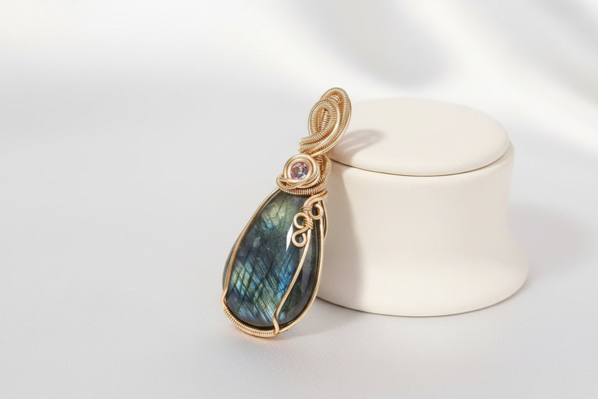 A teardrop-shaped labradorite pendant with a gold wire wrap and a small pink gemstone accent, displayed on a cream ceramic stand.
