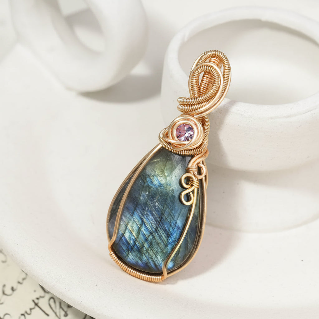 A teardrop-shaped labradorite pendant with a gold wire wrap and a small pink gemstone accent, displayed on a cream ceramic stand.