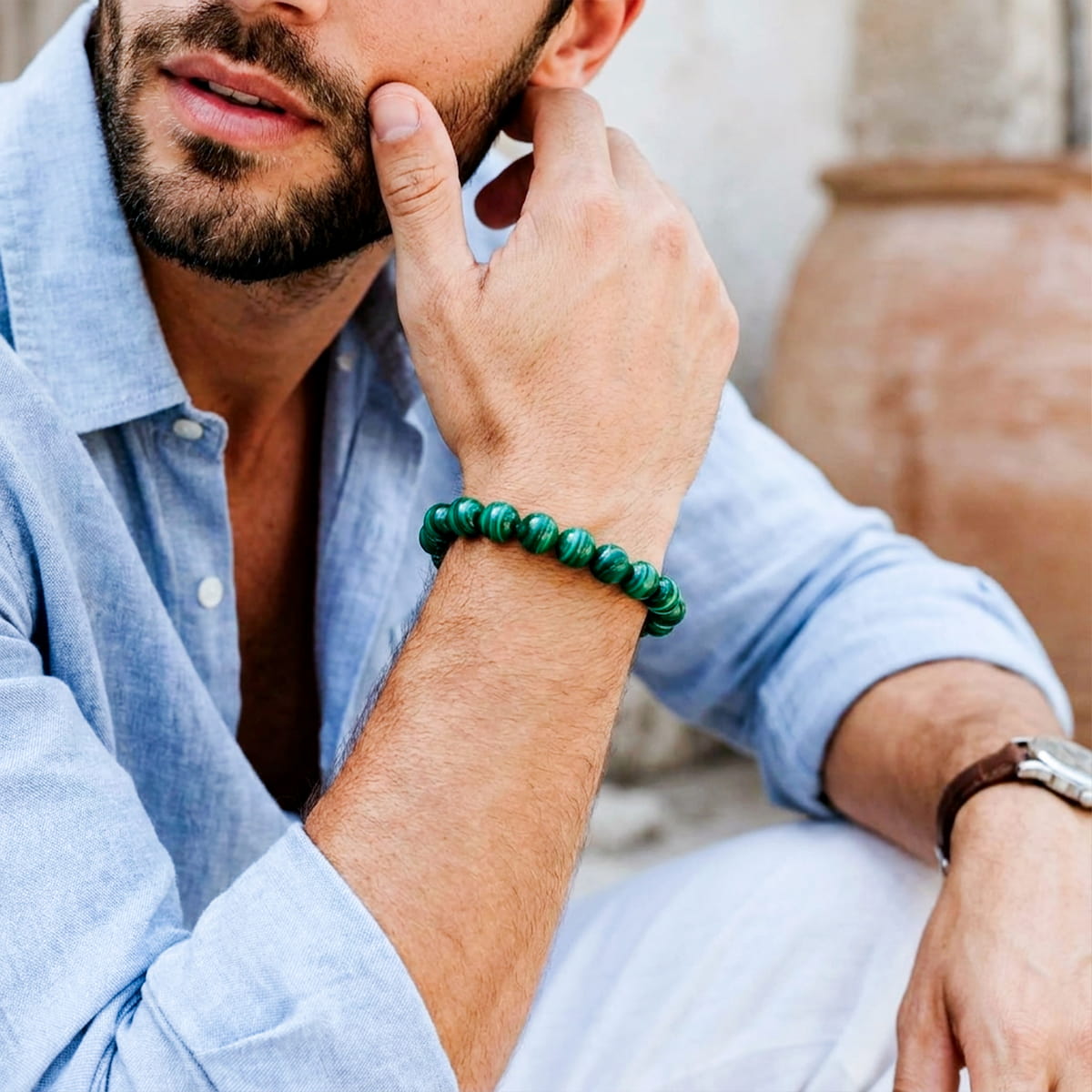 A man wearing a green beaded bracelet on his wrist, wearing a light blue linen shirt and white pants, with a rustic terracotta pot in the background.