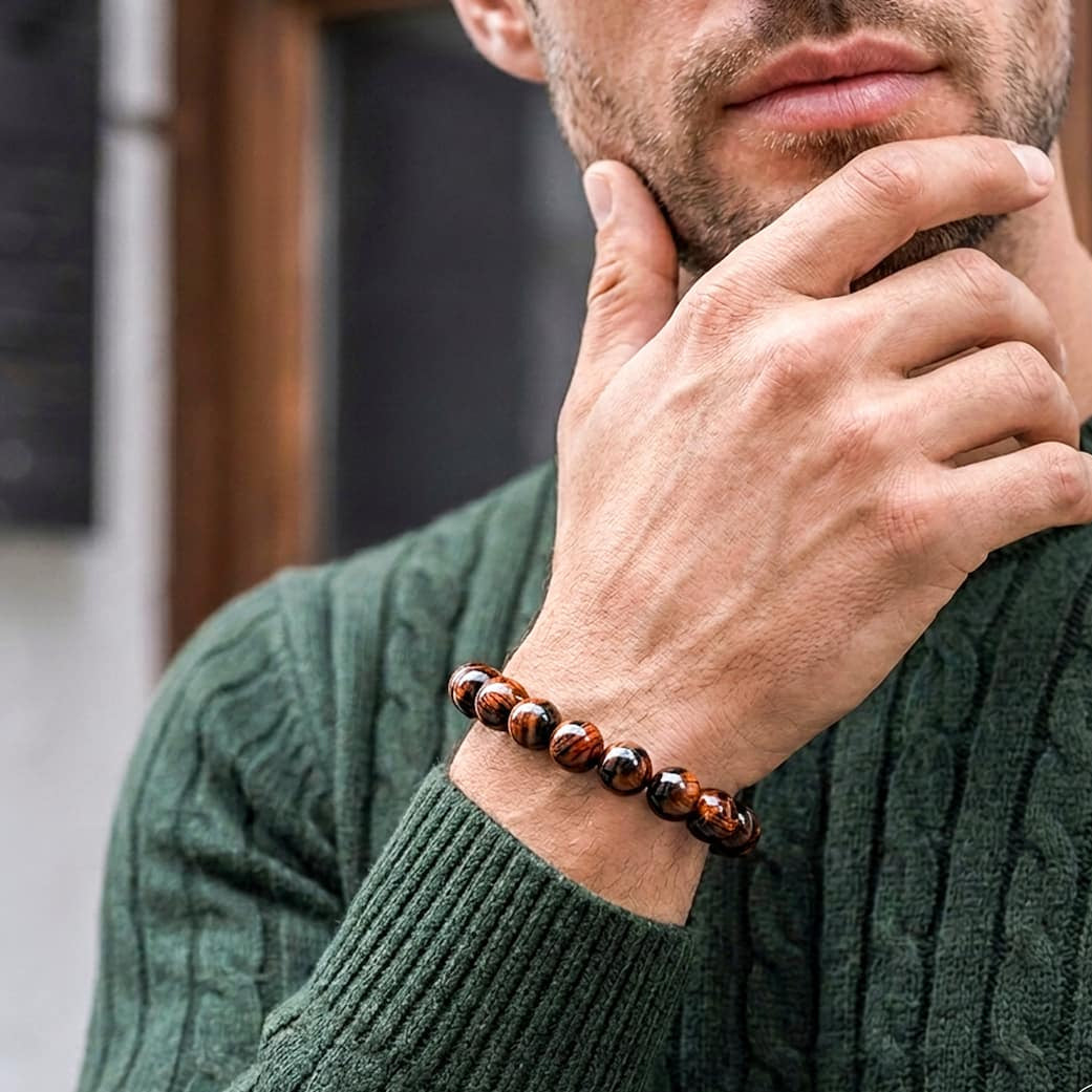 A close-up of a man wearing a dark green cable-knit sweater and a beaded bracelet on his wrist. The bracelet features glossy brown and black tiger's eye beads.