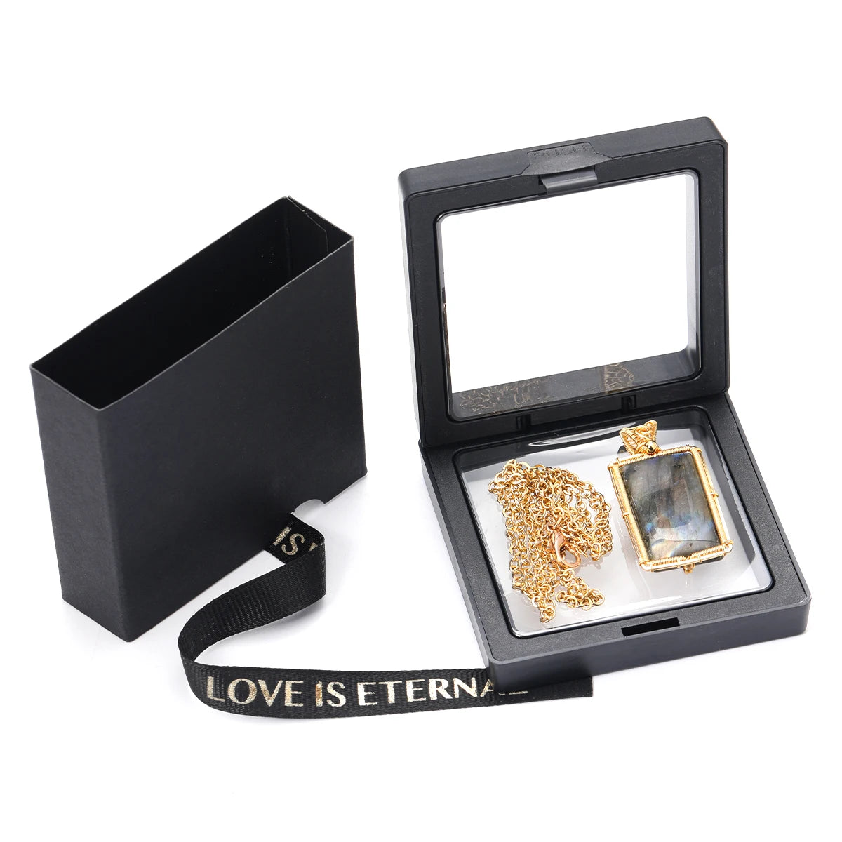 A gold-toned rectangular pendant necklace with a clear glass insert, displayed inside an open black presentation box next to its closed lid. The box has a black ribbon strap with the text 'LOVE IS ETERNA'.
