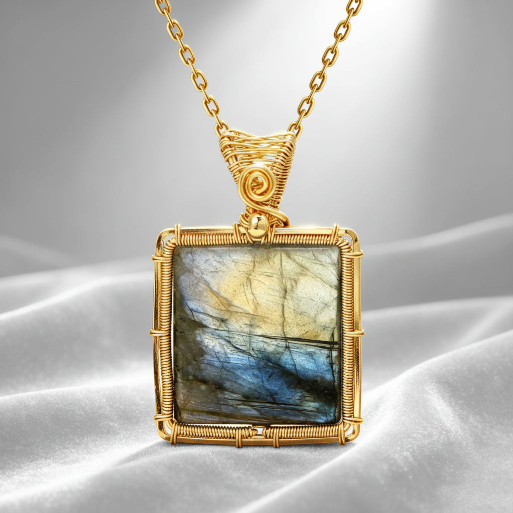 A gold square pendant necklace featuring a polished labradorite stone with shimmering blue, gray, and gold flashes, set in a textured gold wire frame and suspended from a white satin background.