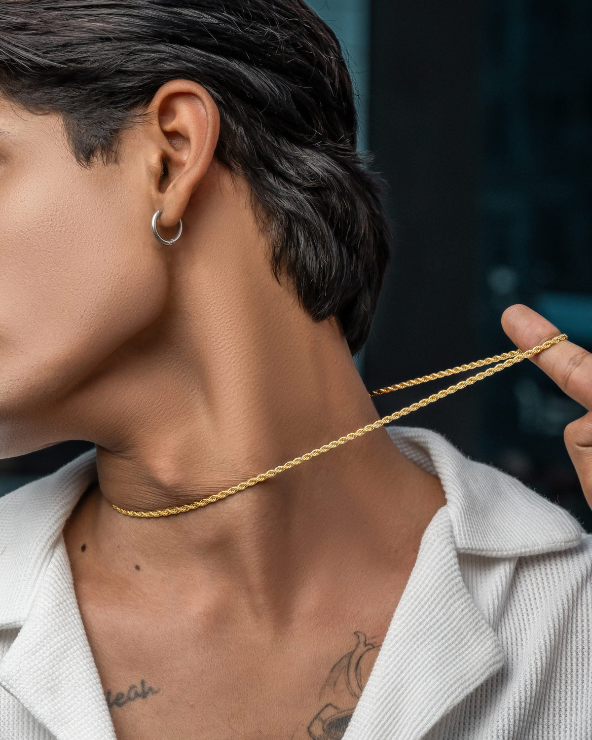 A close-up lifestyle shot of a person wearing a delicate gold chain necklace, holding the ends of the chain with their fingers to show its thinness. The person has short dark hair, a silver hoop earring, and a white textured collared shirt.