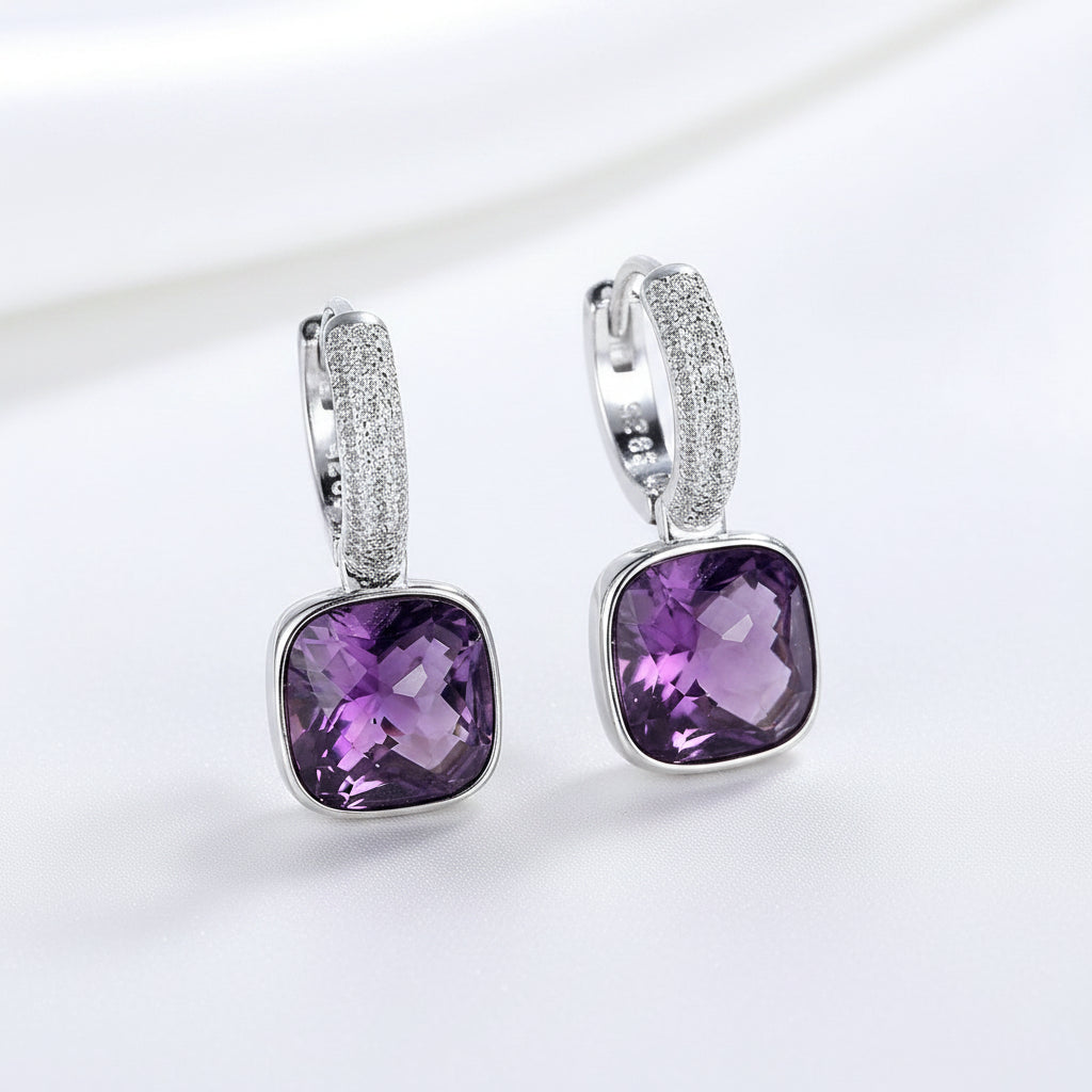 A pair of MEEKCAT sterling silver huggie hoop earrings with square-cut purple amethyst gemstones and pavé-set clear crystals, displayed on a light blue textured surface with a '925 Real Sterling' stamp overlay.