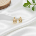 A pair of gold-toned stud earrings with raw, irregular crystal formations, displayed on a white textured surface next to a wood slice and green leaves. 