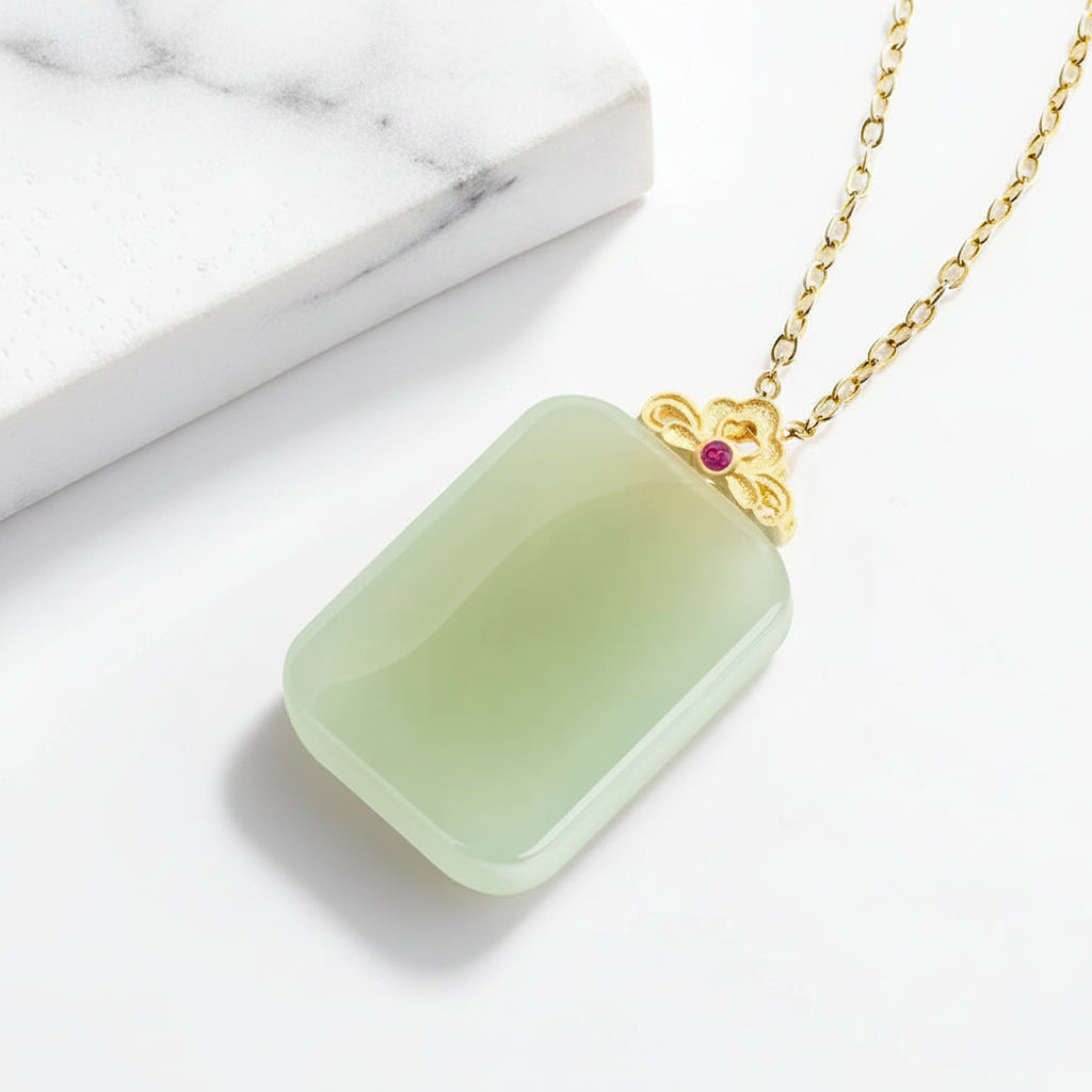 A rectangular light green jade pendant necklace with a gold-tone floral clasp and a small red gemstone, displayed on a white surface with a white book prop. The brand logo 'Mengfancy' is visible in the top left corner.