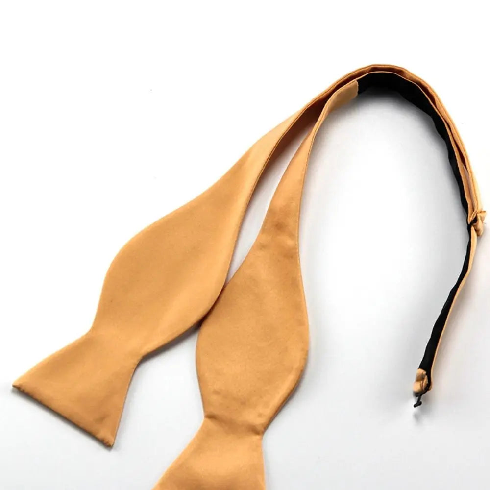 A pre-tied bow tie in a warm camel tan color, laid flat on a clean white background. The inner lining is black, visible along the curved strap.
