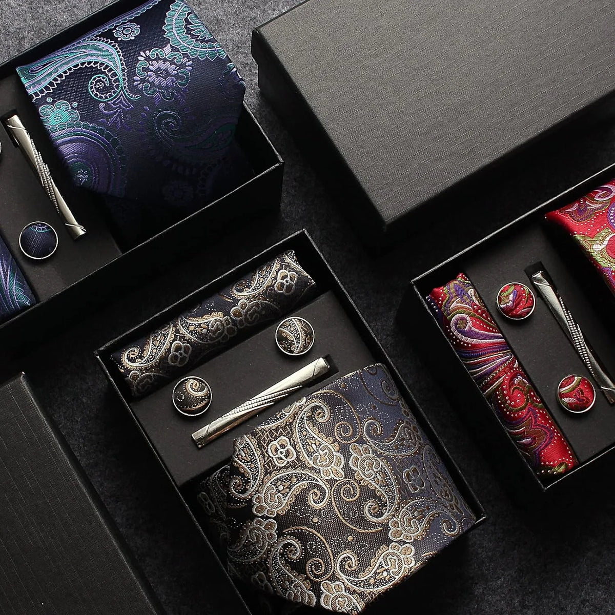 Set of paisley patterned tie, pocket square, and accessories in black boxes on a dark surface.