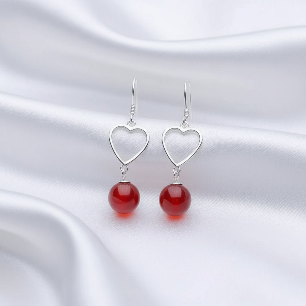 A pair of silver heart-shaped dangle earrings with smooth, glossy red bead drops, displayed on a white jewelry stand against a dark background.