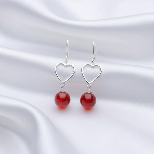 A pair of silver heart-shaped dangle earrings with smooth, glossy red bead drops, displayed on a white jewelry stand against a dark background.