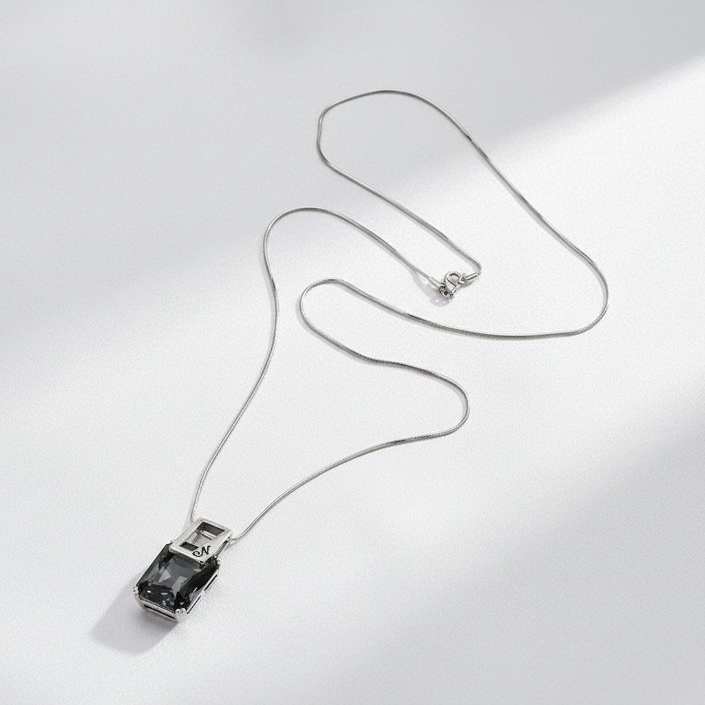 A silver-toned snake chain necklace with a rectangular pendant featuring a dark smoky purple stone, displayed on a textured  white background