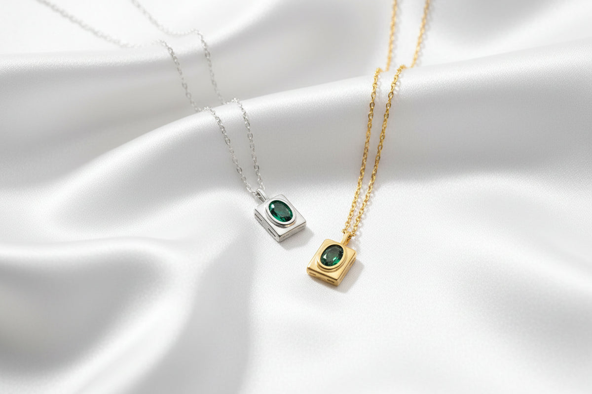 Two sterling silver square pendant necklaces with oval green gemstones, one in a silver frame and one in a gold frame, displayed on chains against a silky white background 