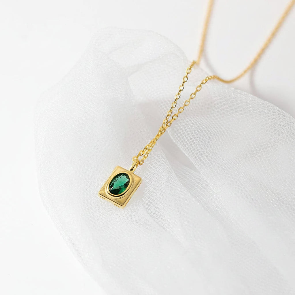 A gold necklace with a square bezel setting holding a green oval gemstone, displayed on white textured fabric. A red circular stamp in the lower right reads 'REAL STERLING 925'.
