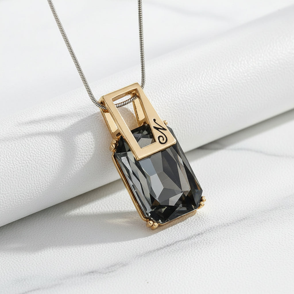 A silver-toned rectangular pendant necklace with a faceted dark smoky crystal and a script letter 'M' on the top plate, shown on a textured white background.