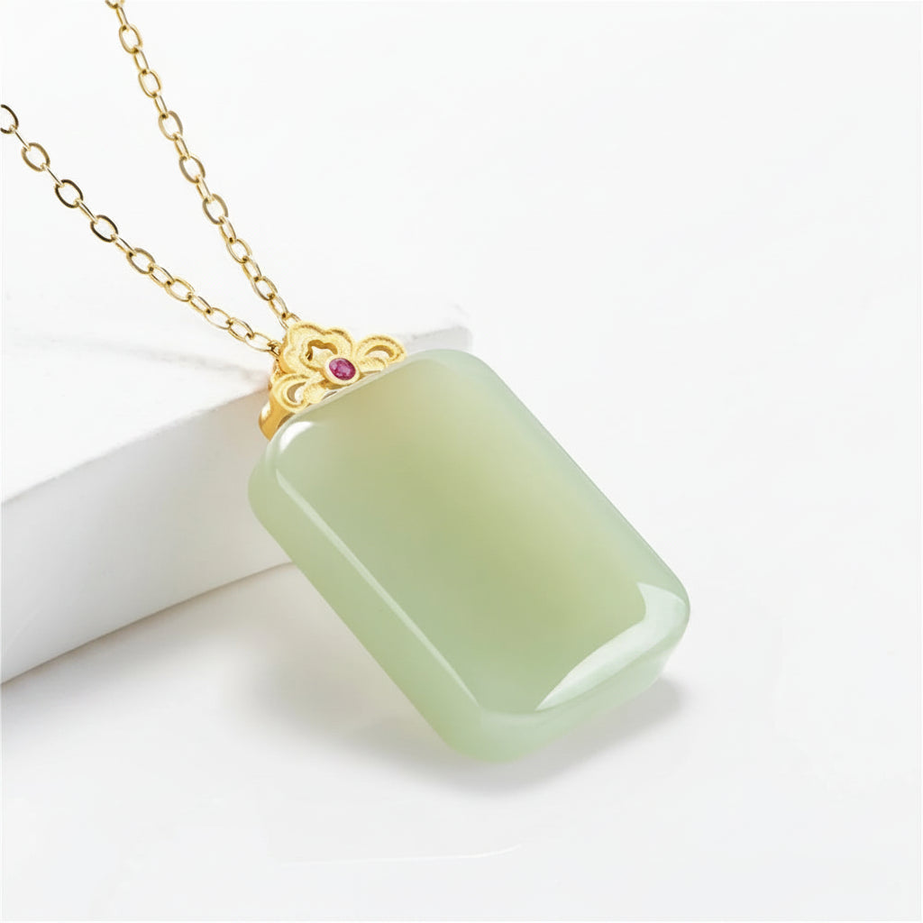 A rectangular light green jade pendant necklace with a gold-tone chain and decorative gold cap featuring a small pink gemstone, displayed on a white block against a white background.