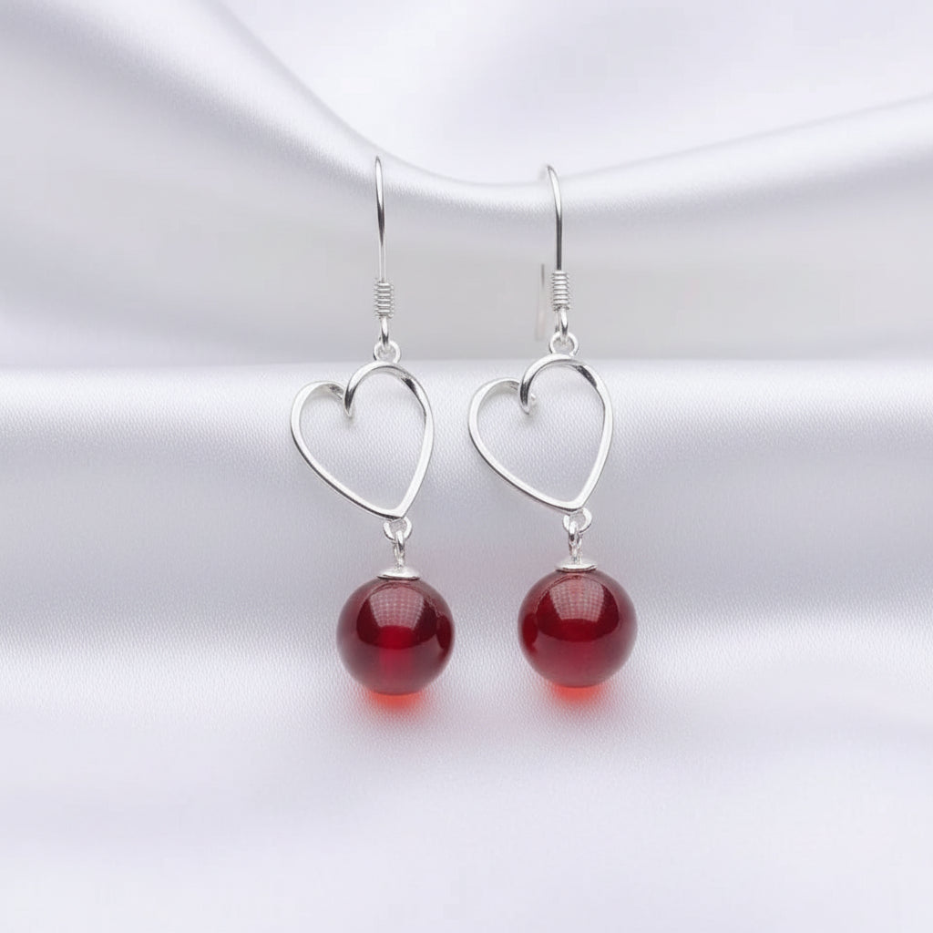 A pair of silver heart dangle earrings with red bead drops, displayed on a white jewelry stand against a black background.