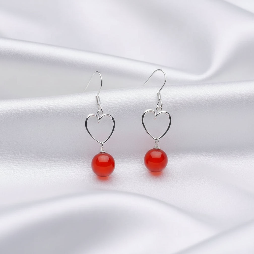 A pair of silver heart-shaped dangle earrings with smooth, glossy red spherical beads, displayed on a white surface against a black background.