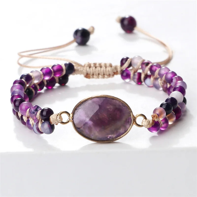 A beaded stretch bracelet featuring a large oval purple amethyst stone in a gold-toned bezel, flanked by strands of round purple, violet, and dark maroon beads with a tan cord closure.