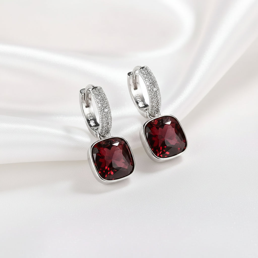 A pair of MEEKCAT sterling silver huggie hoop earrings with square-cut dark red gemstones and a curved row of clear crystals. The earrings are displayed on a white surface with a white ceramic dish and a gray faux fur texture in the background, featuring a 'REAL STERLING 925' stamp in the corner.