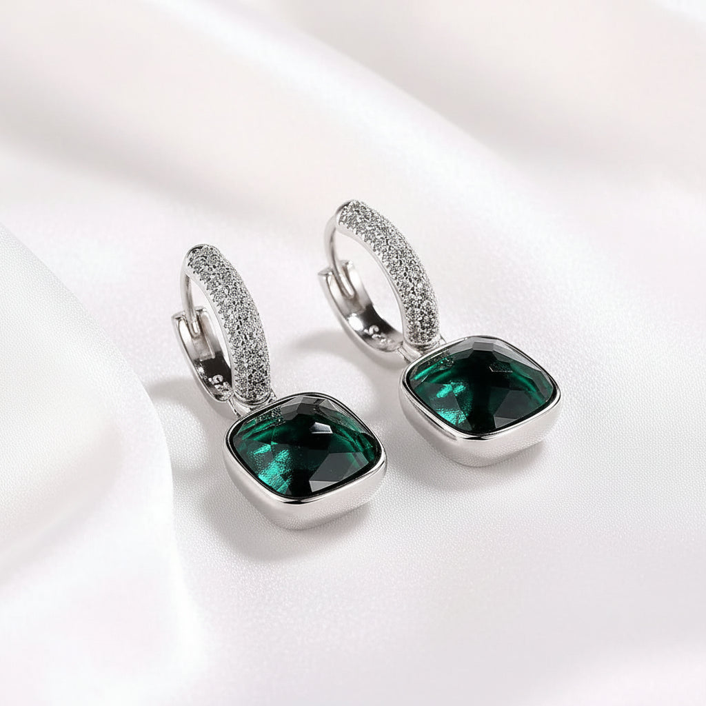 A pair of MEEKCAT silver huggie hoop earrings with square-cut green gemstones and a band of small clear crystals, displayed on a white textured surface with a paper background and a 'REAL STERLING 925' stamp.