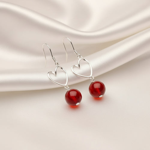 A pair of silver dangle earrings with open heart-shaped wire frames and smooth, glossy red bead drops, displayed on a white rectangular stand against a black background.