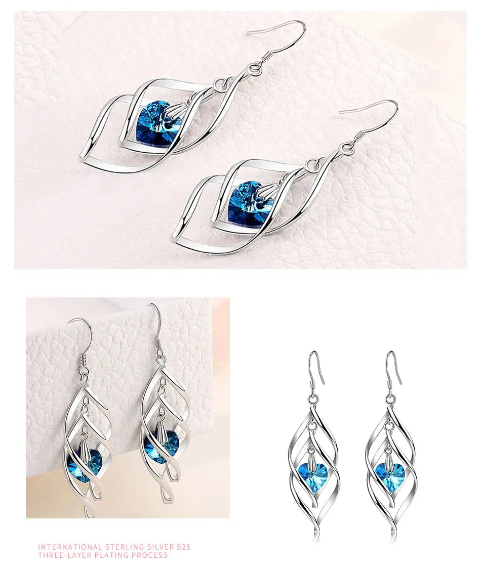 Three views of silver dangle earrings with a blue heart-shaped crystal in a leaf-like layered setting, shown on a light textured background.