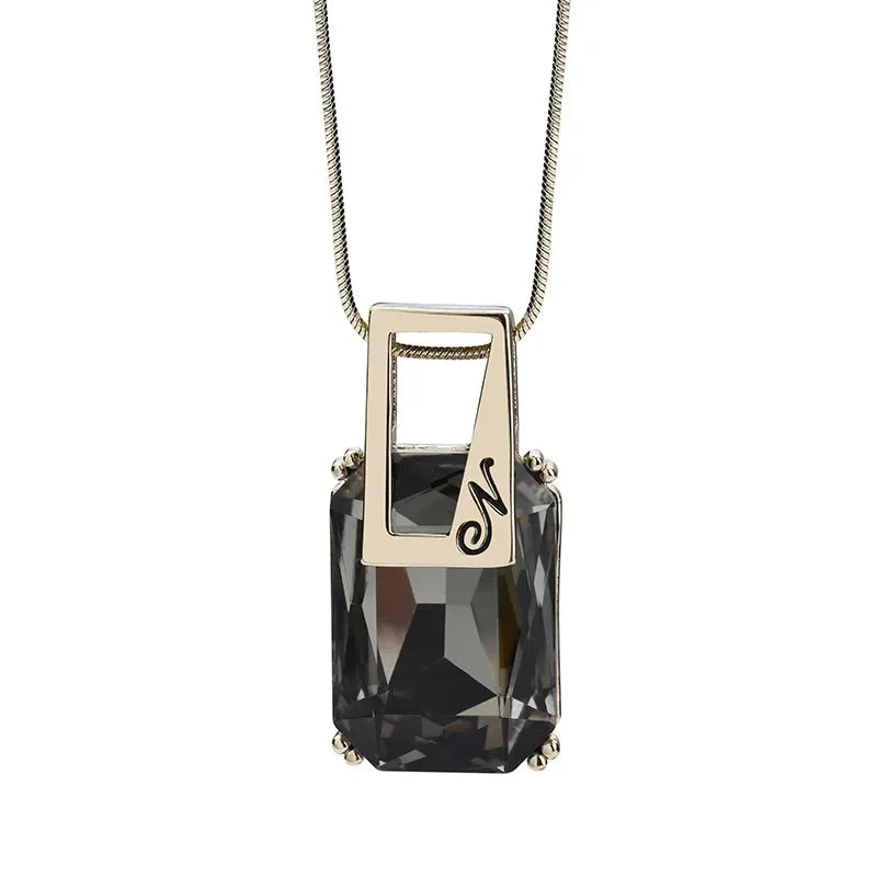 A rectangular faceted dark gray smoky crystal pendant necklace with a polished silver-toned metal frame and chain, featuring an engraved script letter 'N' on the side.