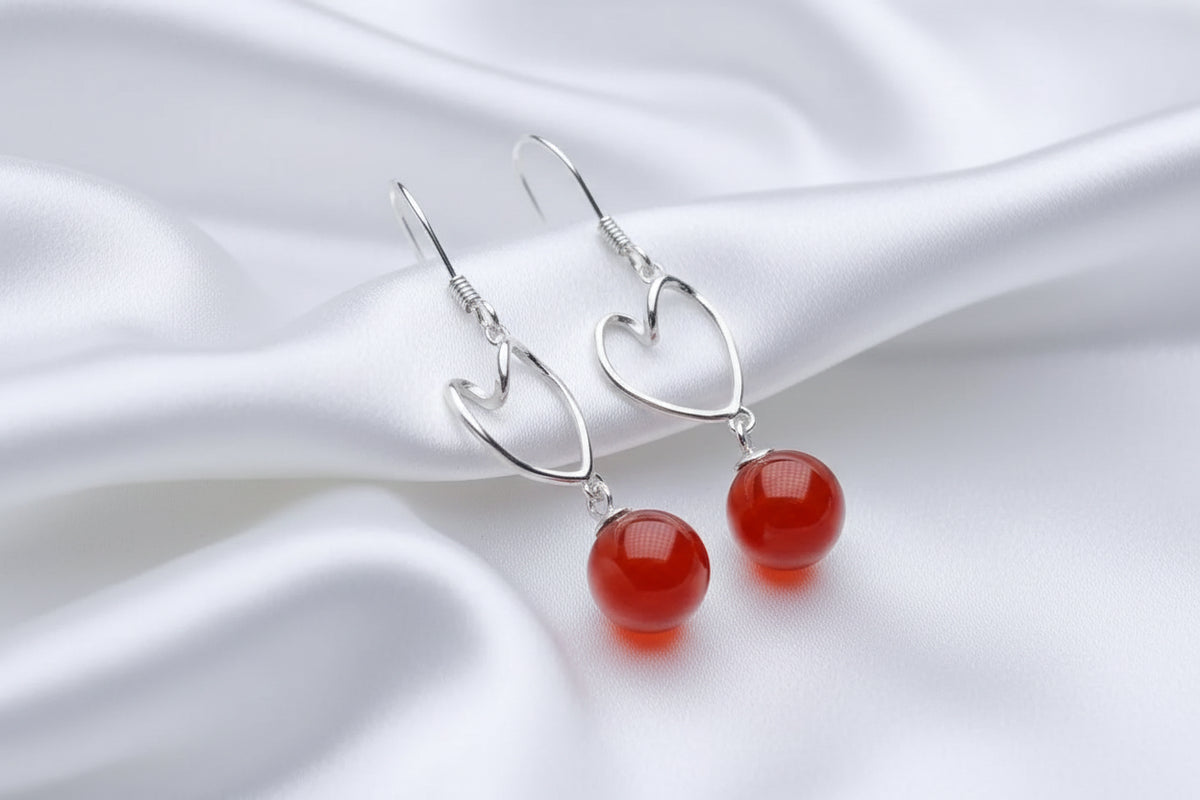 A pair of silver drop earrings with smooth, round red gemstone beads, displayed on a white rectangular surface against a black background.