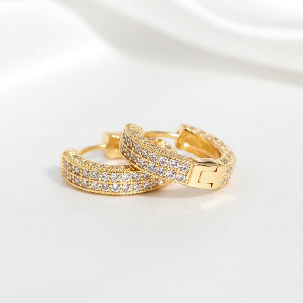 Two gold-toned bracelets adorned with clear pavé stones, displayed on a reflective gray surface with a sparkling overlay effect. 