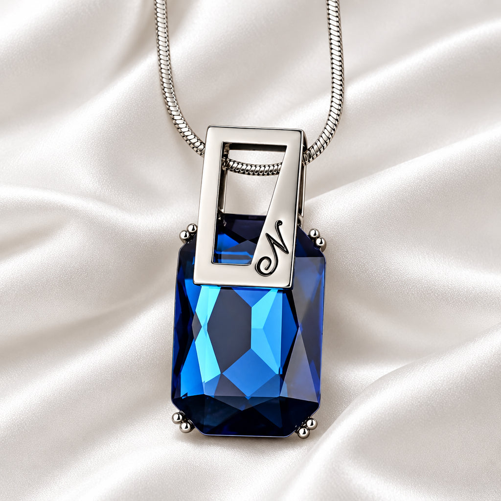 A silver pendant necklace featuring a faceted rectangular blue gemstone and a polished silver frame engraved with a script letter 'N', suspended from a silver chain against a white background.
