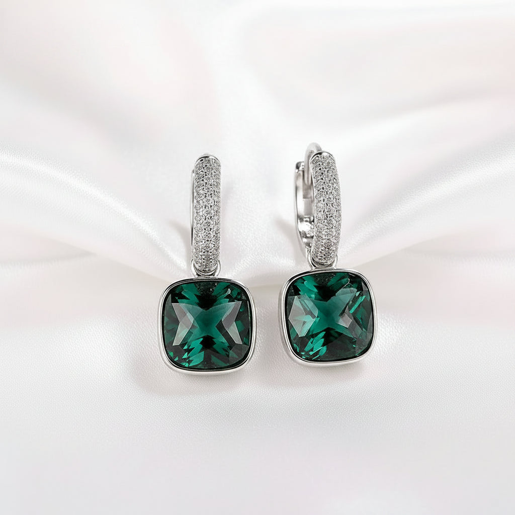 A pair of MEEKCAT silver huggie hoop earrings with square-cut green gemstones and a band of small clear crystals, displayed on a white textured surface with a paper background and a 'REAL STERLING 925' stamp.