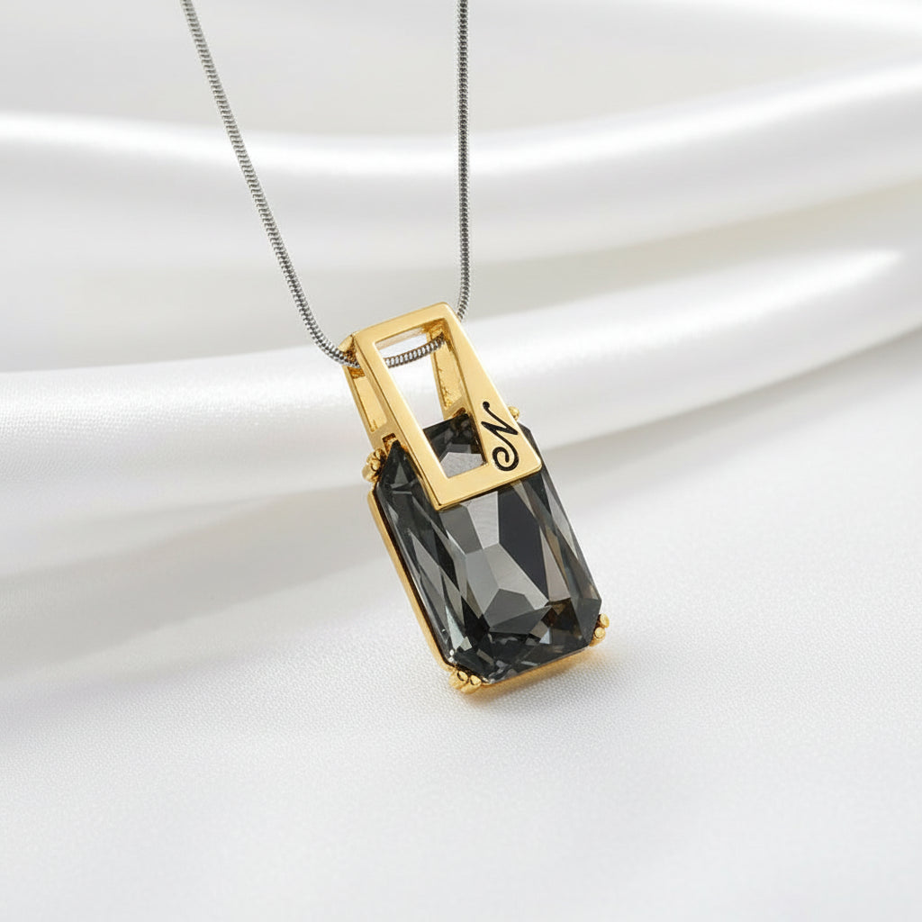 A rectangular faceted dark gray gemstone pendant on a silver-toned chain, displayed on a textured white background