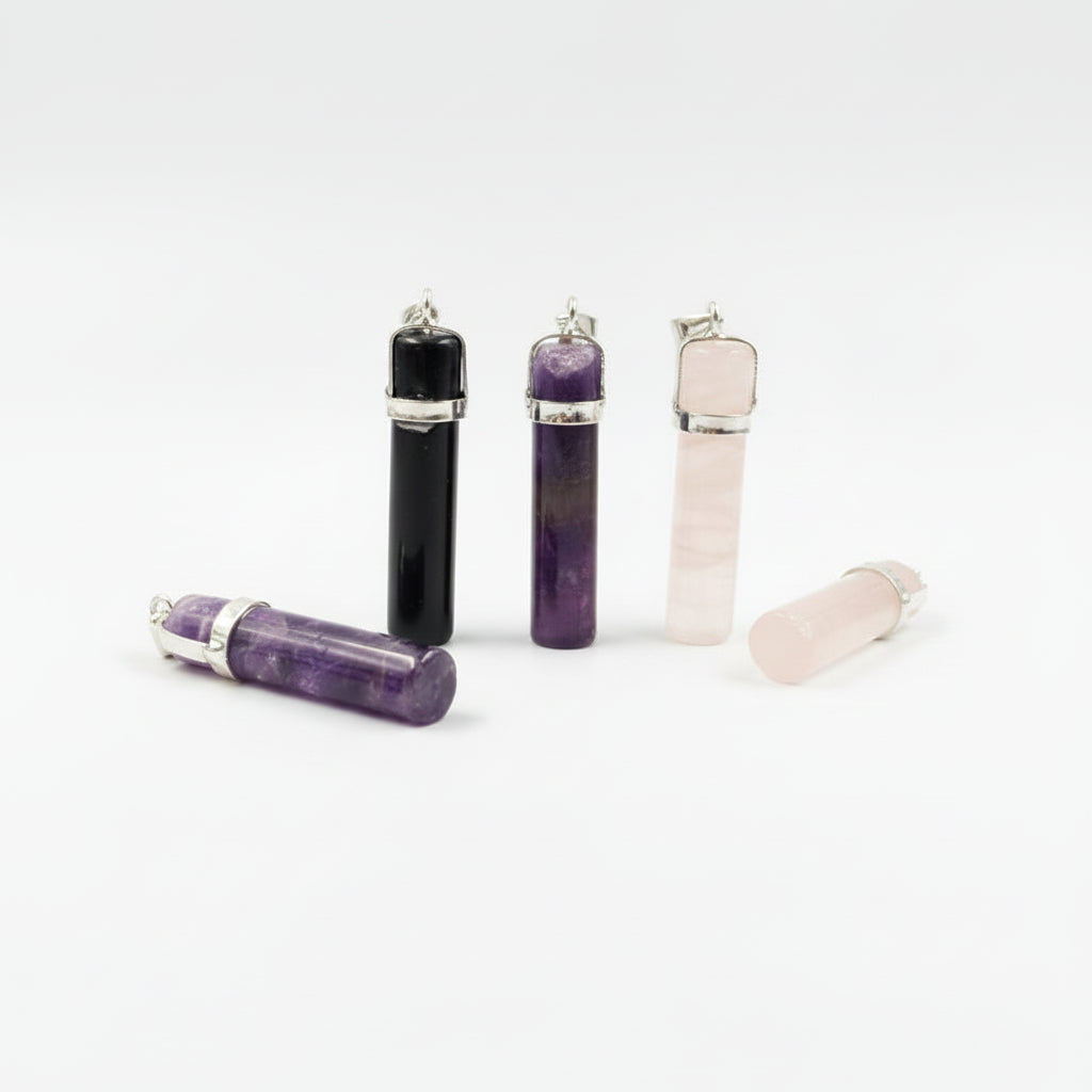 Four cylindrical crystal pendants or oil diffuser necklaces displayed on a gray background, including black, purple amethyst, pale pink quartz, and a loose purple crystal with silver-toned caps.