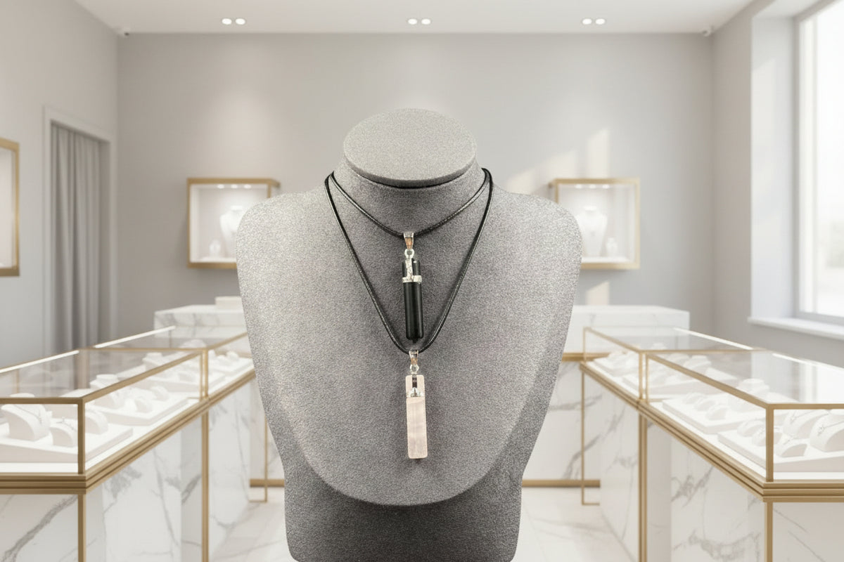 A black cord necklace featuring a dual pendant design: a vertical black cylindrical pendant with silver-toned caps and a larger rectangular clear stone pendant hanging below, displayed on a gray mannequin bust against a white background.