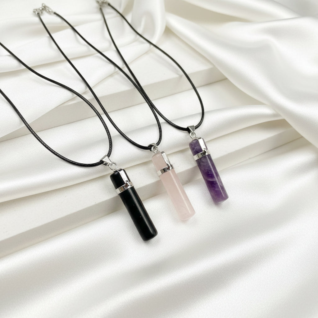 Three cylindrical crystal pendant necklaces on black cords, displayed diagonally on a textured dark gray surface. The necklaces feature polished black, pale pink, and purple crystals with silver metal caps.