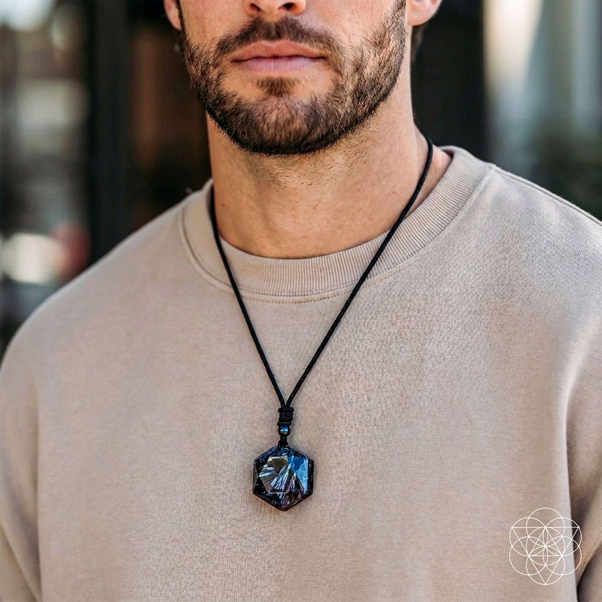 A close-up of a man wearing a black cord necklace with a faceted dark blue and black pendant, styled with a beige crewneck sweatshirt.