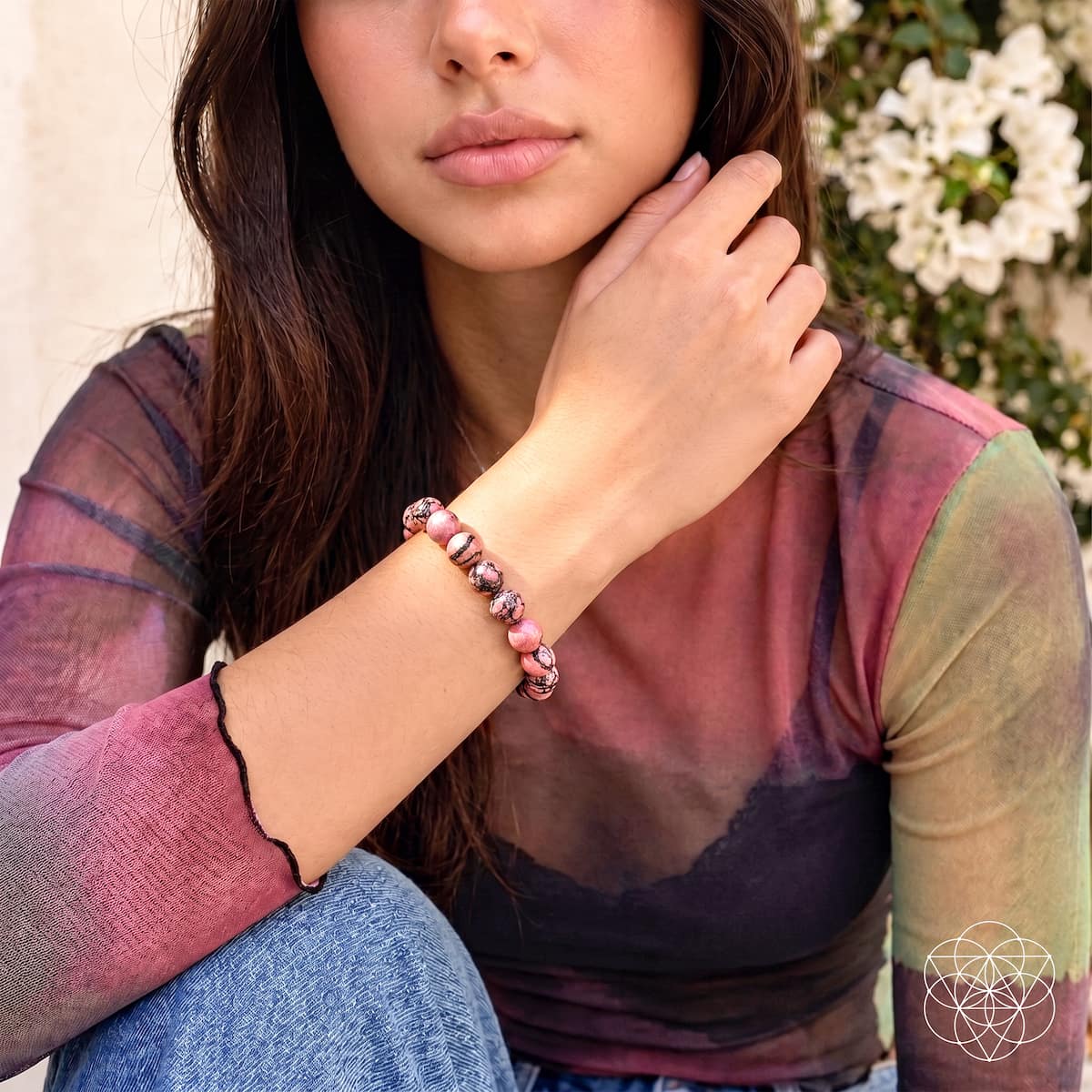 A woman wearing a pink and brown beaded bracelet on her wrist, wearing a sheer long-sleeve top with a pink and green gradient, sitting in front of a soft-focus background of white flowers.