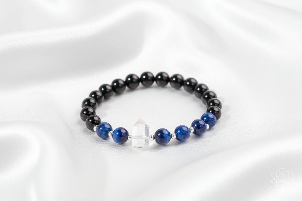 A beaded bracelet with glossy black round beads, deep blue lapis lazuli-style beads, a clear faceted crystal charm, and silver spacer beads, arranged on a white silky background.