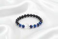 A beaded bracelet with glossy black round beads, deep blue lapis lazuli-style beads, a clear faceted crystal charm, and silver spacer beads, arranged on a white silky background.